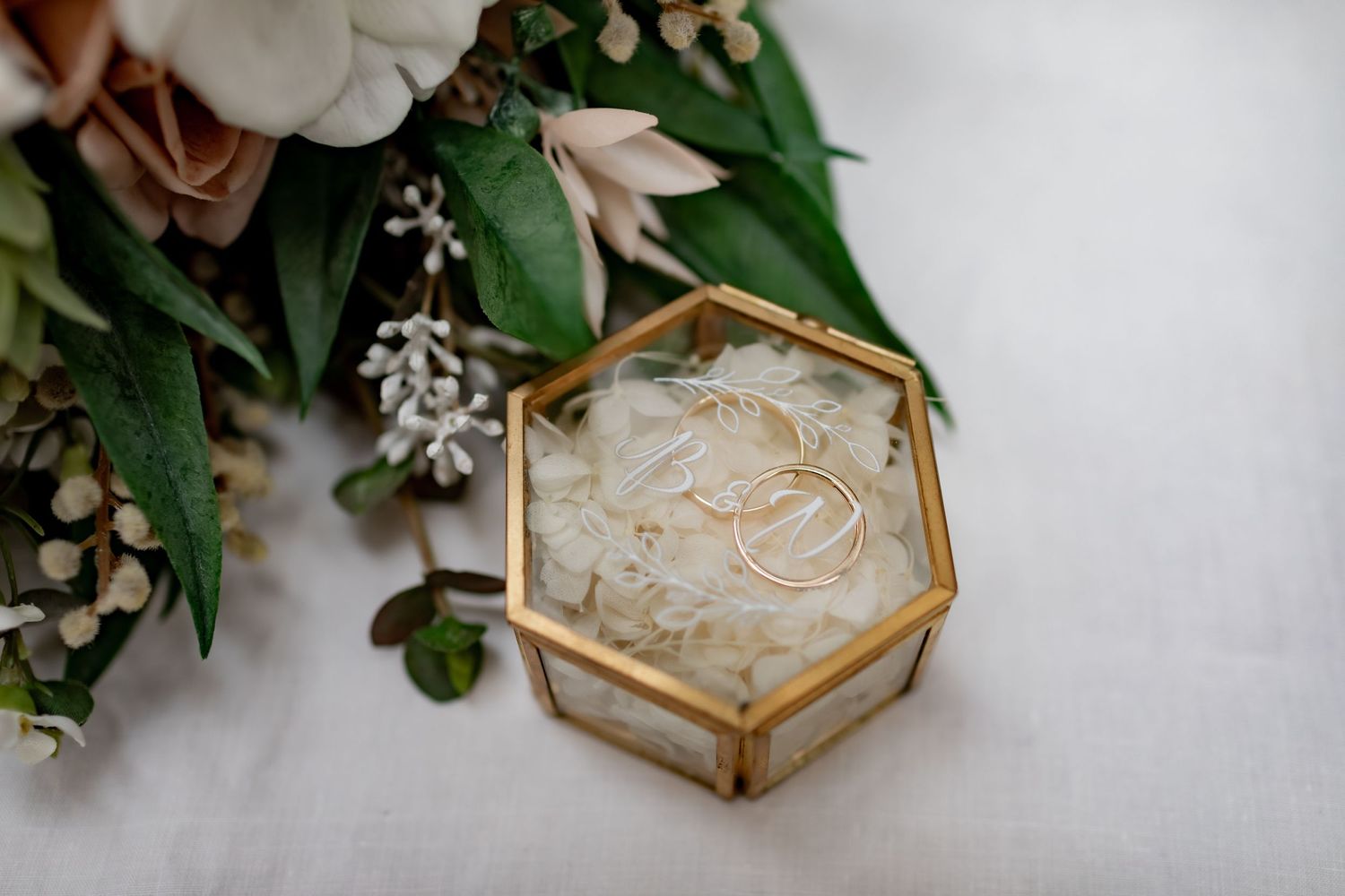 Wedding rings sit in geometric glass box surrounded by floral arrangements and greenery.