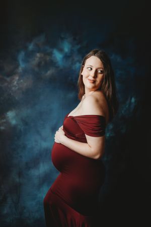Artistic maternity photos in burgundy dress against dramatic blue-tinted background.