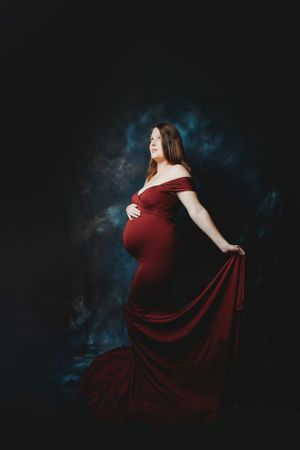 Flowing burgundy maternity gown creates dramatic silhouette against dark backdrop.