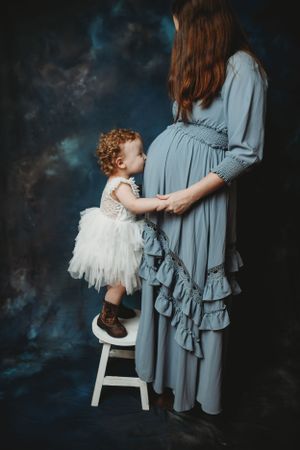 Child in white dress poses near figure in flowing gray dress against dark backdrop.