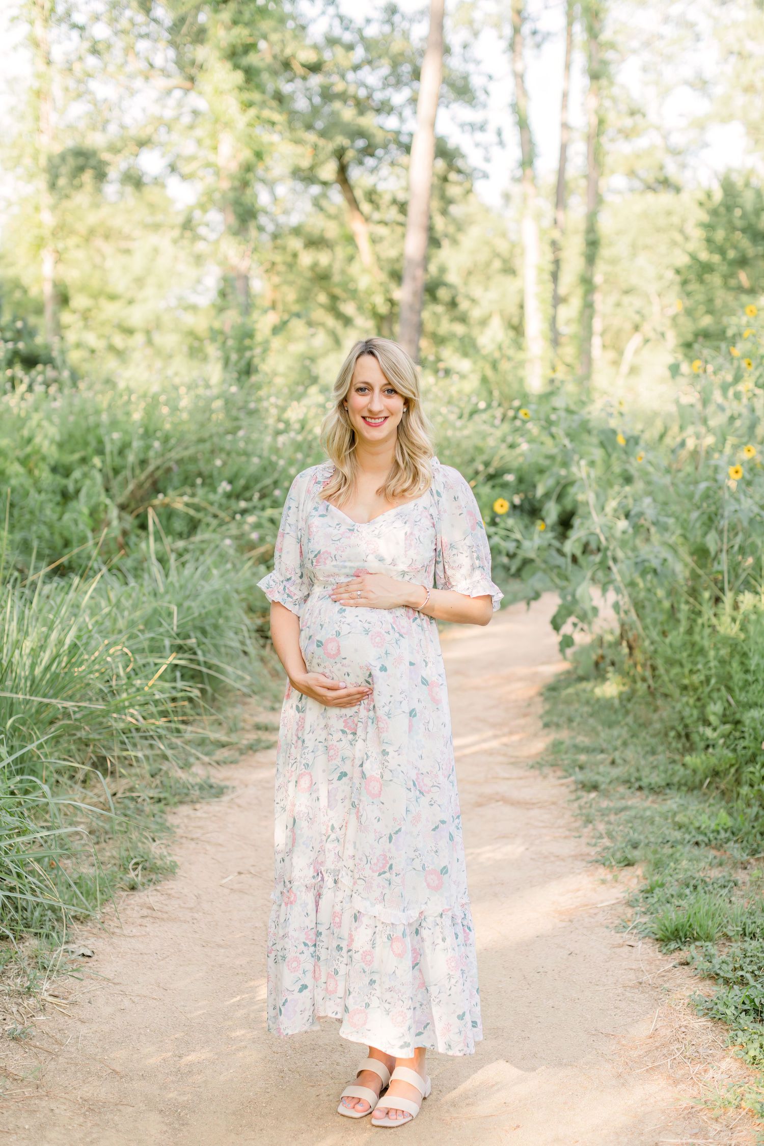 Sun-drenched maternity photo featuring flowing floral maxi dress on garden path.