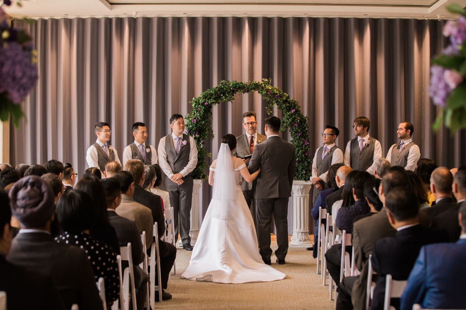 Indoor wedding ceremony under simple greenery arch with guests seated in rows.