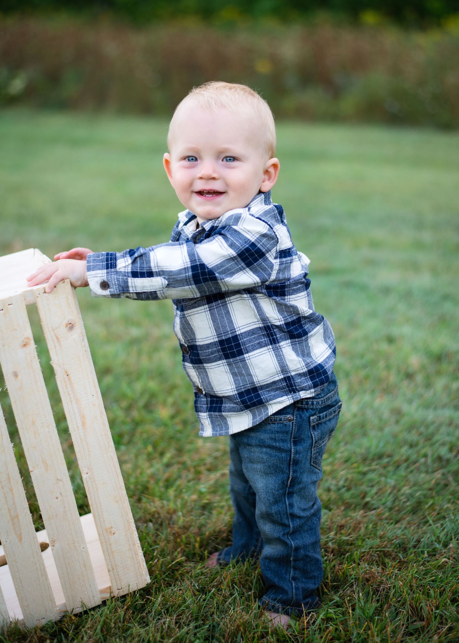 A toddler in plaid flannel and jeans smiles while holding onto a wooden crate outdoors.