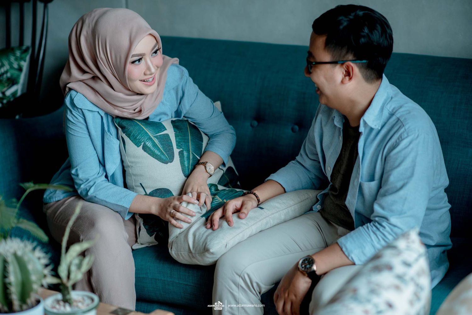 A couple engages in conversation while sitting on a teal couch surrounded by houseplants.