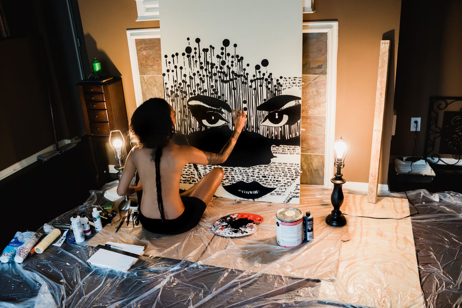 An artist paints a black and white floral mural on a white wall while sitting on a protected floor.