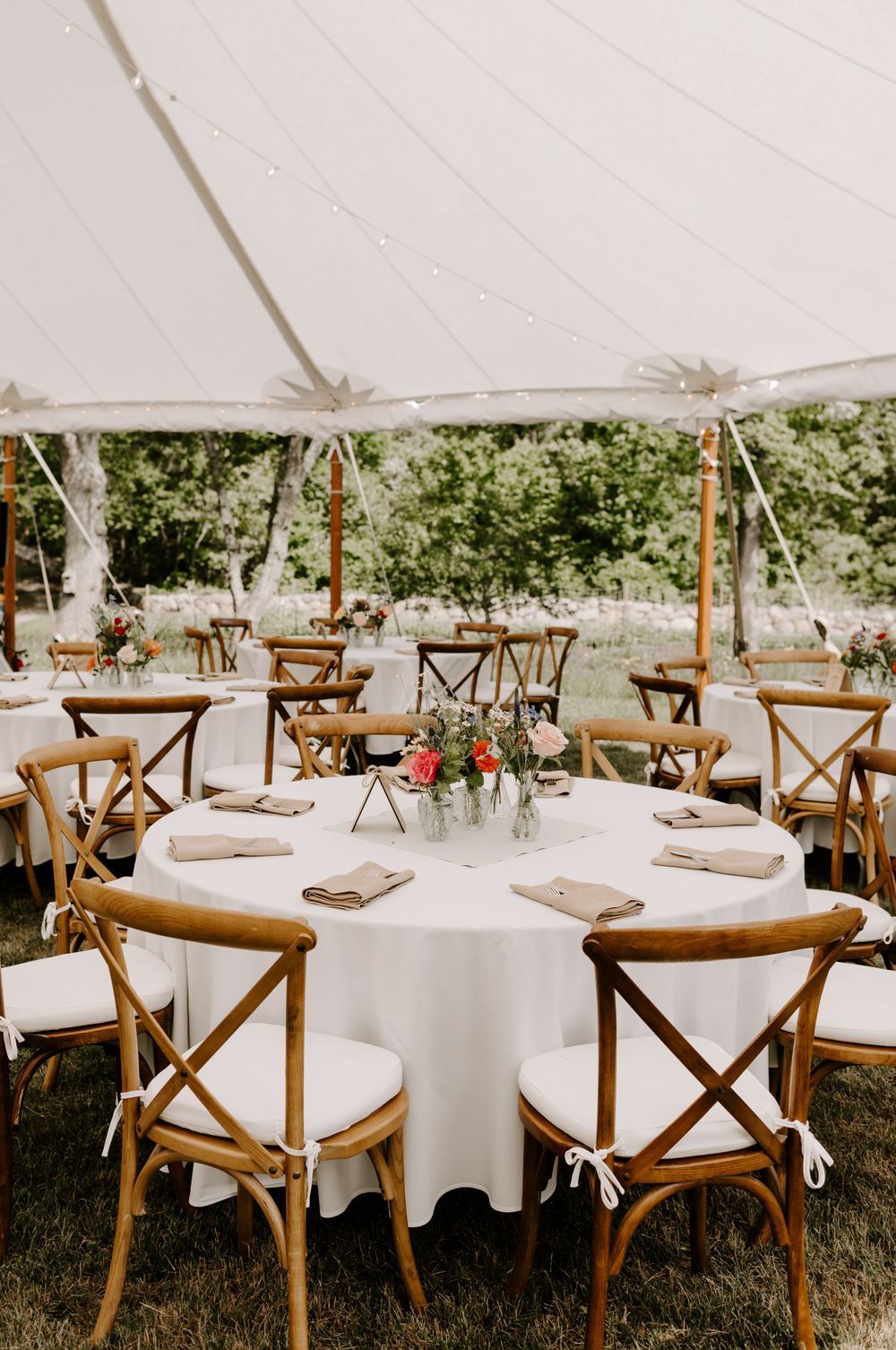 Elegant outdoor wedding reception with white tablecloths and cross-back wooden chairs under a tent.