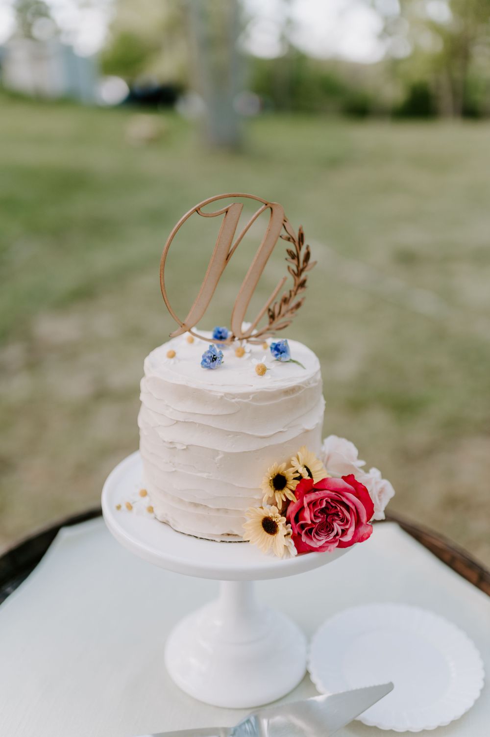 Small white wedding cake with gold monogram topper and pink rose decoration on a white pedestal stand.