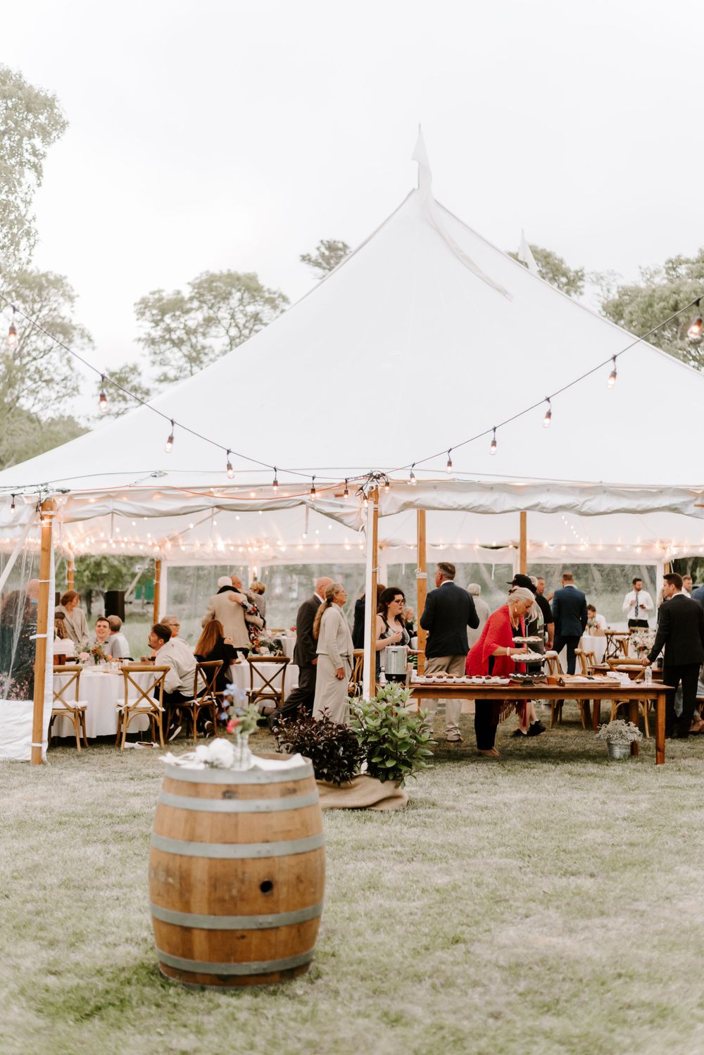 White tent wedding reception setup with wine barrels and string lights in a garden setting.