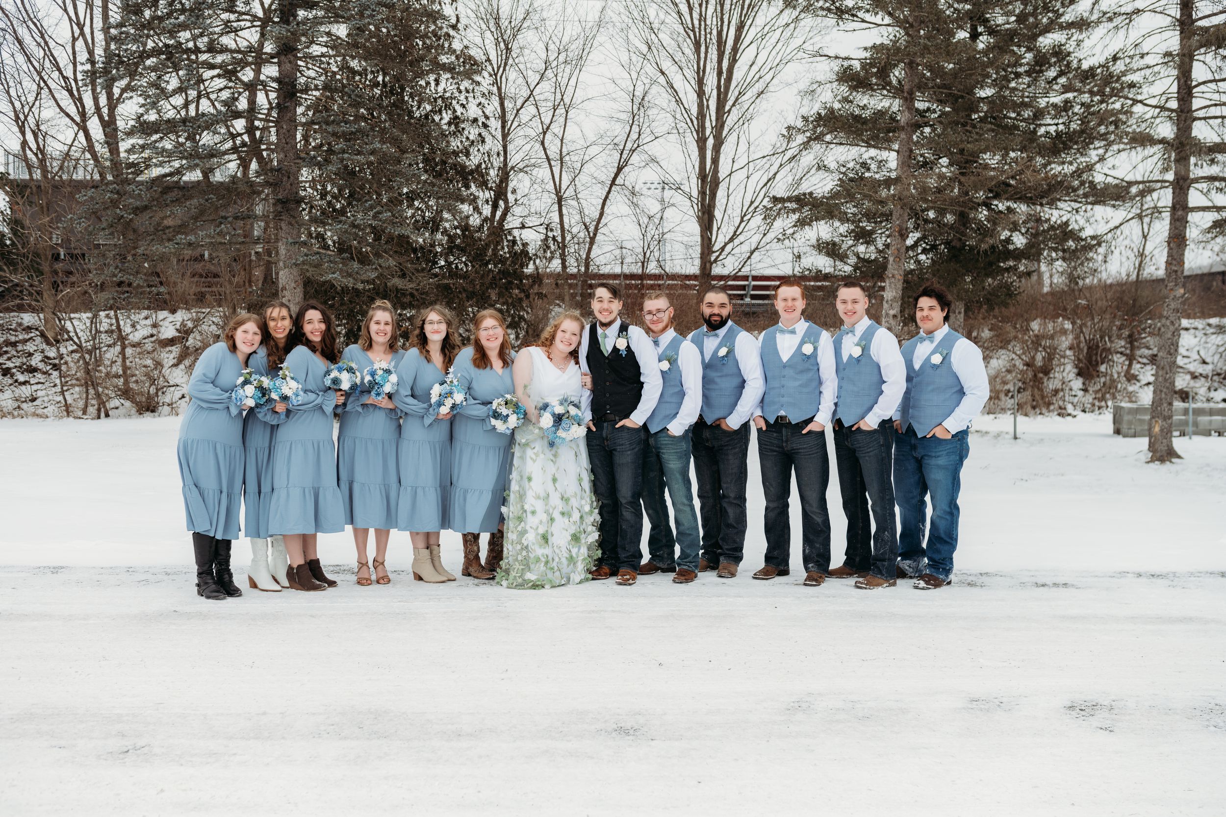 Wedding party poses in light blue attire against snowy winter backdrop with evergreen trees.
