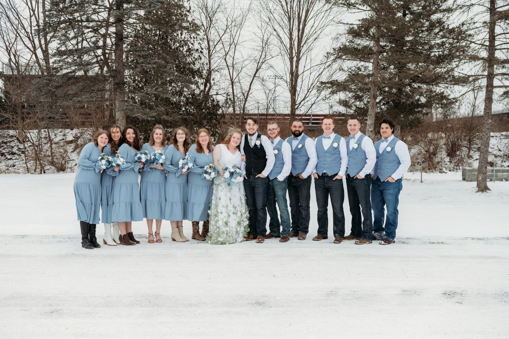 Wedding party poses in light blue attire against snowy winter backdrop with evergreen trees.