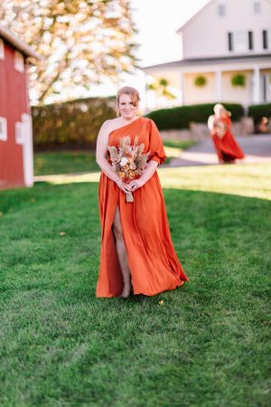 Bridesmaid walks across lawn in vibrant orange gown holding autumn-toned bouquet.