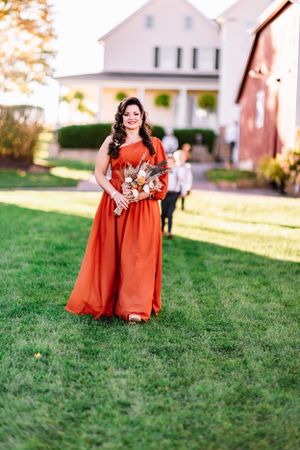 Woman in flowing burnt orange dress holds a bouquet against white farmhouse backdrop.