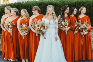 Bride in white lace gown poses with bridesmaids wearing flowing burnt orange dresses holding autumn bouquets.