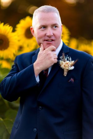 Groom in navy suit with boutonniere poses thoughtfully against backdrop of sunflowers.