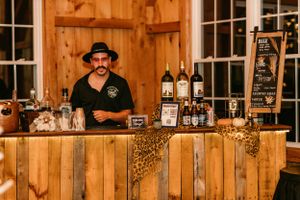 A bartender stands behind a rustic wooden bar counter with bottles and glassware on display.