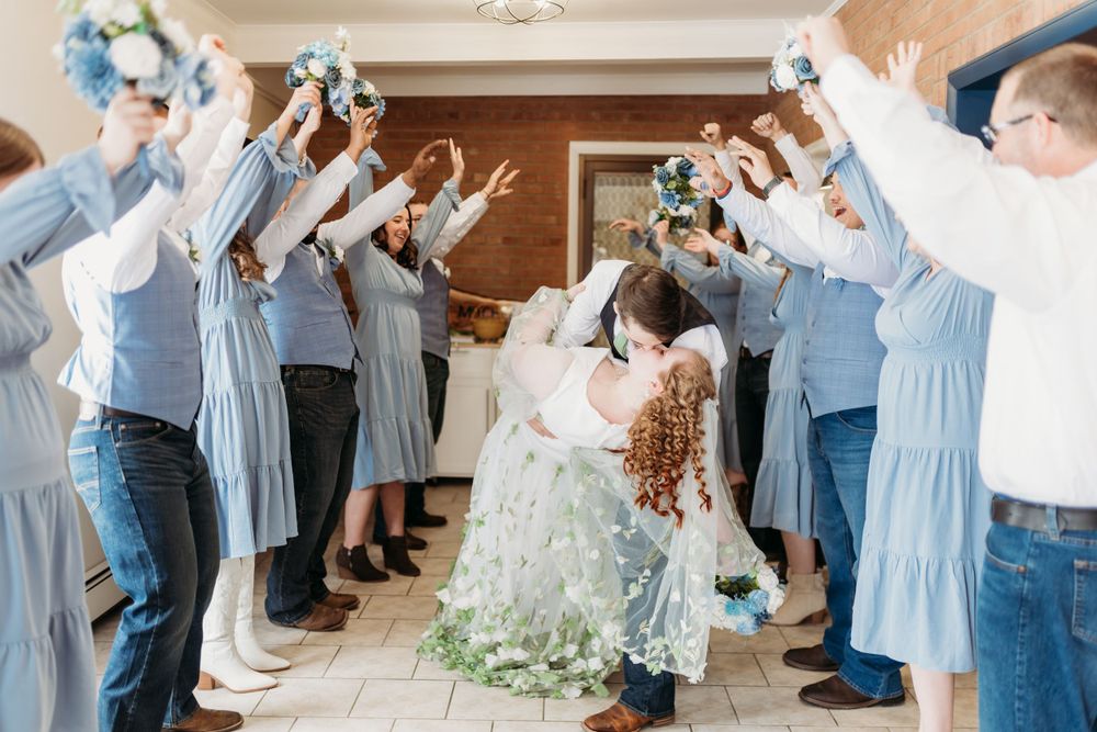 Wedding party celebrates in blue attire as couple poses under an arch indoors.