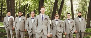 Groomsmen in light gray suits with bowties standing together in a wooded outdoor setting.