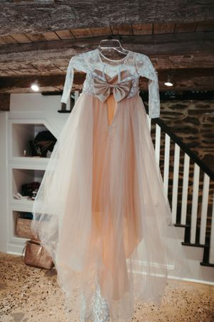 A delicate flower girl dress with bow detail hanging against rustic wooden beams.