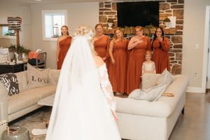 Bridesmaids in rust-colored dresses gathered around a displayed wedding gown in a modern living room.