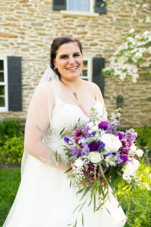 Bride in white dress holds vibrant purple and white floral bouquet against stone building backdrop.