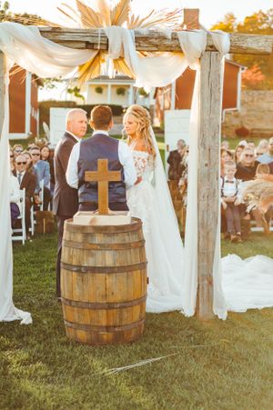 Outdoor wedding ceremony with wooden barrel altar and white fabric canopy against red barn backdrop.