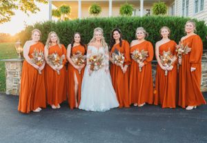 Bridal party in bright orange dresses poses at sunset with greenery backdrop.