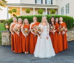 Bridal party in vibrant orange dresses stands in front of white colonial house with hanging ferns.