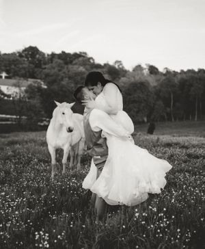 Black and white wedding photo of a couple sharing an intimate moment with a white horse in a meadow.