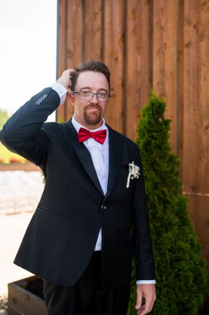 A groom in a black tuxedo and red bowtie adjusts his hair outside a wooden barn.