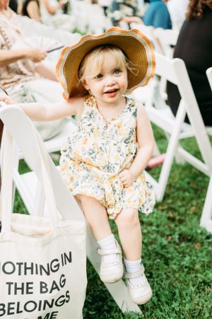 Small child in floral romper and sun hat sitting on white chair at outdoor event.