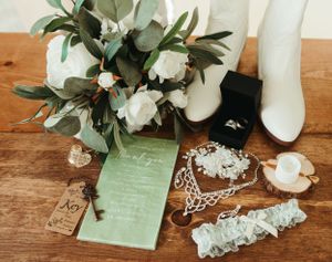 Wedding details including white boots, floral bouquet, invitation and lace garter on wooden surface.