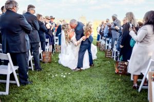 Wedding guests gather around a central aisle with white chairs and scattered flower petals during an outdoor ceremony.