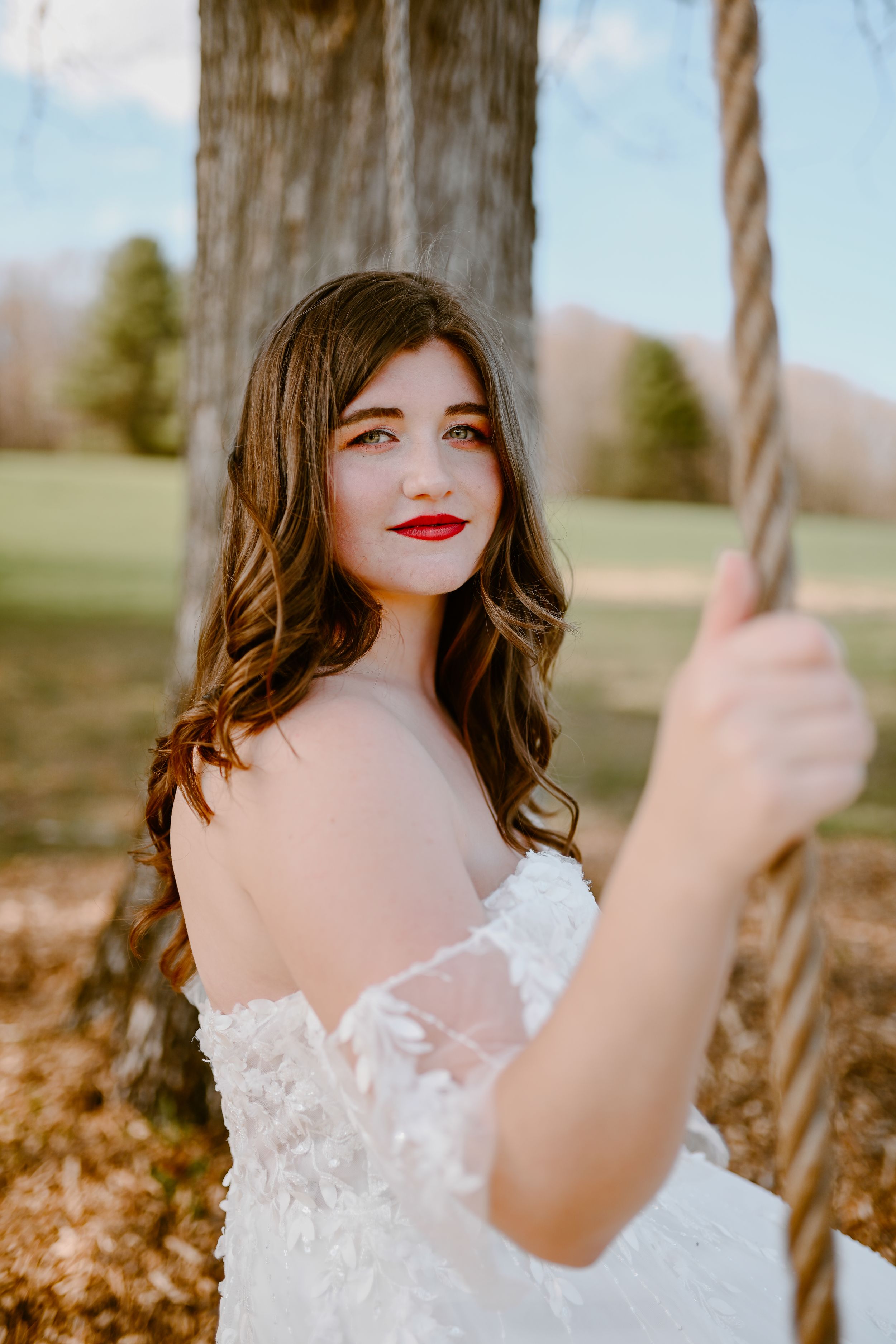 A bride in lace dress poses outdoors near a wooden swing on an autumn day.