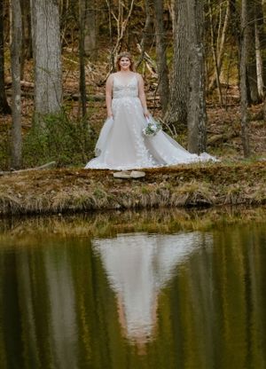 A bride in a white gown stands by a reflective pond in a wooded forest setting.