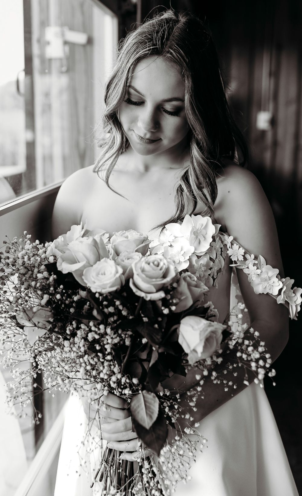 A bride in white dress admires her bouquet of roses and baby's breath in black and white.