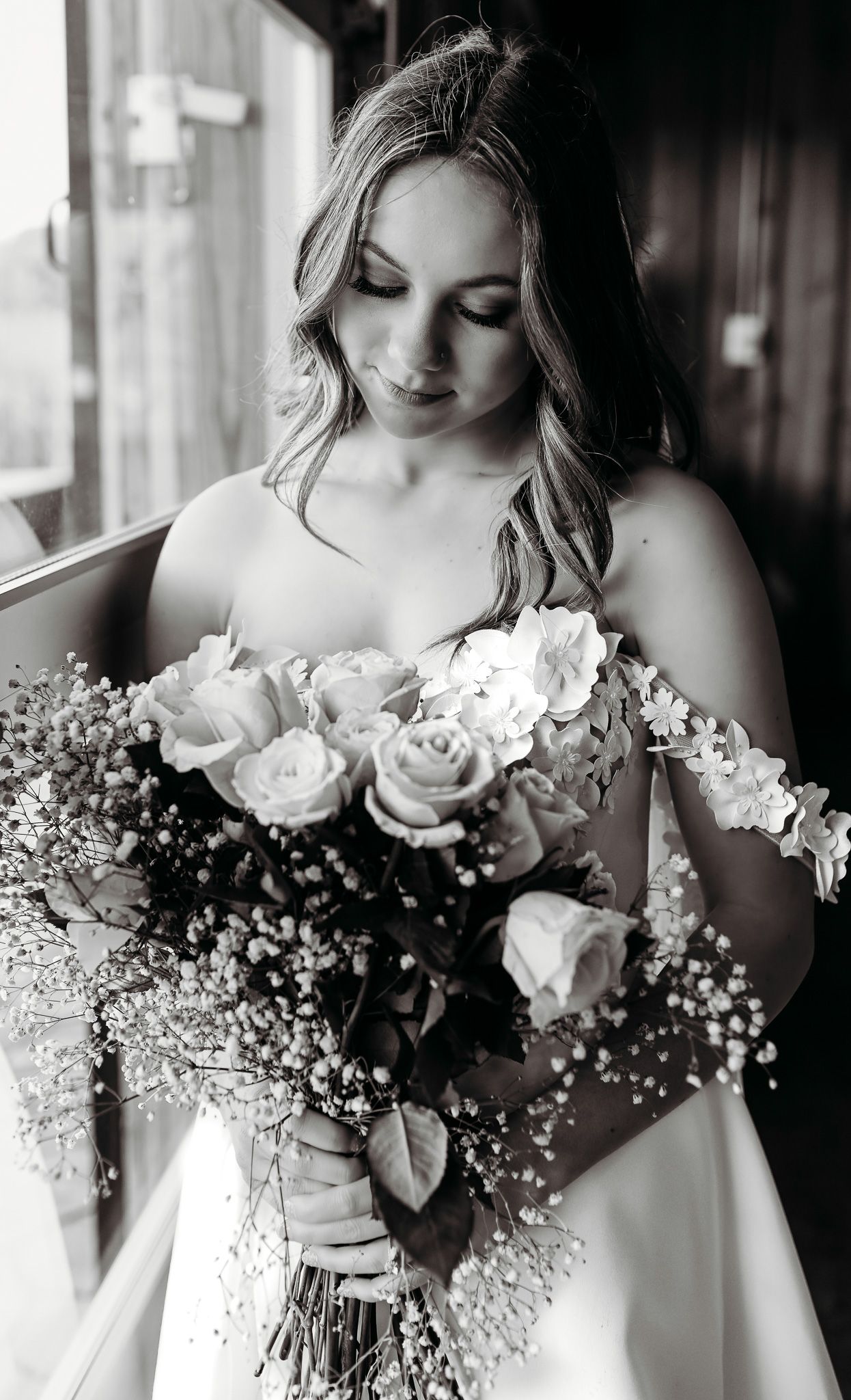A bride in white dress admires her bouquet of roses and baby's breath in black and white.