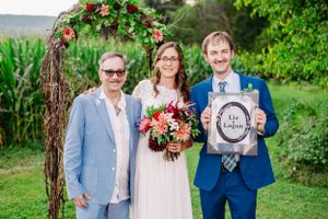 Wedding party poses under rustic floral arch with decorative sign in garden setting.