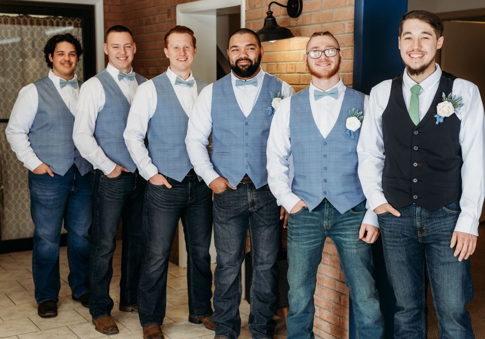 Groomsmen lined up wearing matching grey vests and dress shirts against a brick wall.