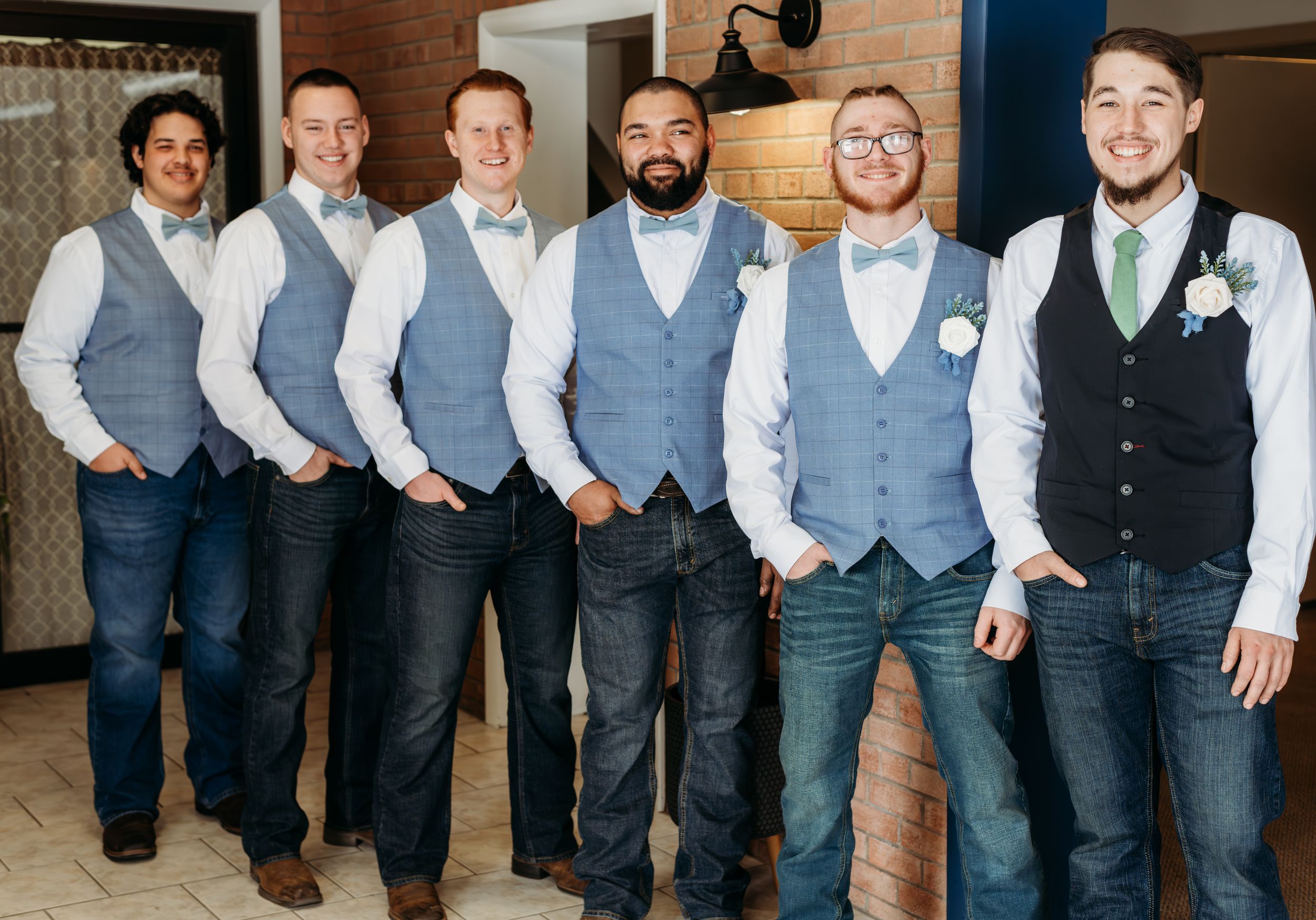 Groomsmen lined up wearing matching grey vests and dress shirts against a brick wall.