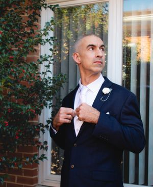 A groom in a navy suit adjusts his tie while standing by a window.