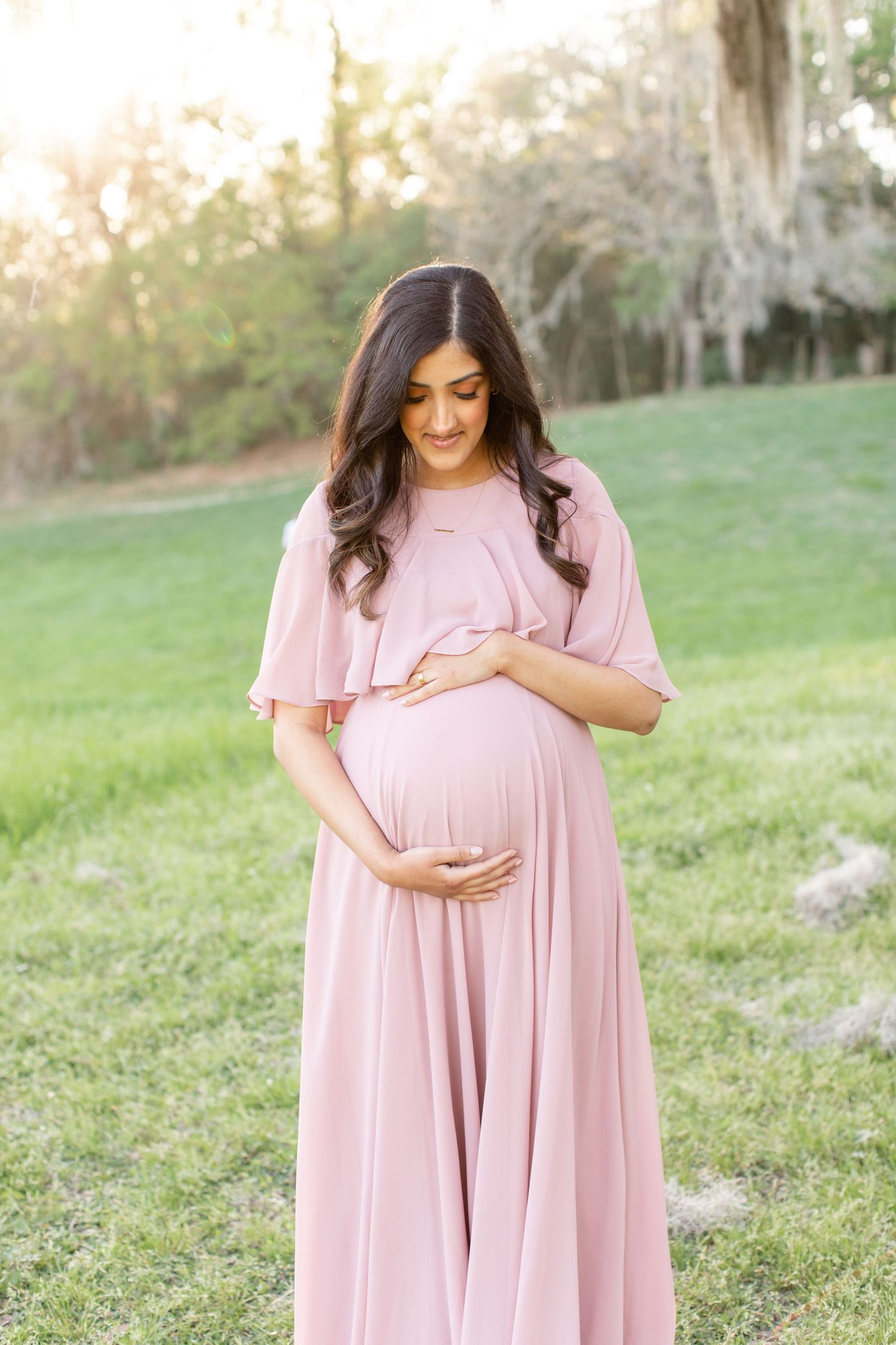 Pregnant woman in pink dress tenderly holds baby bump while standing in natural outdoor setting.