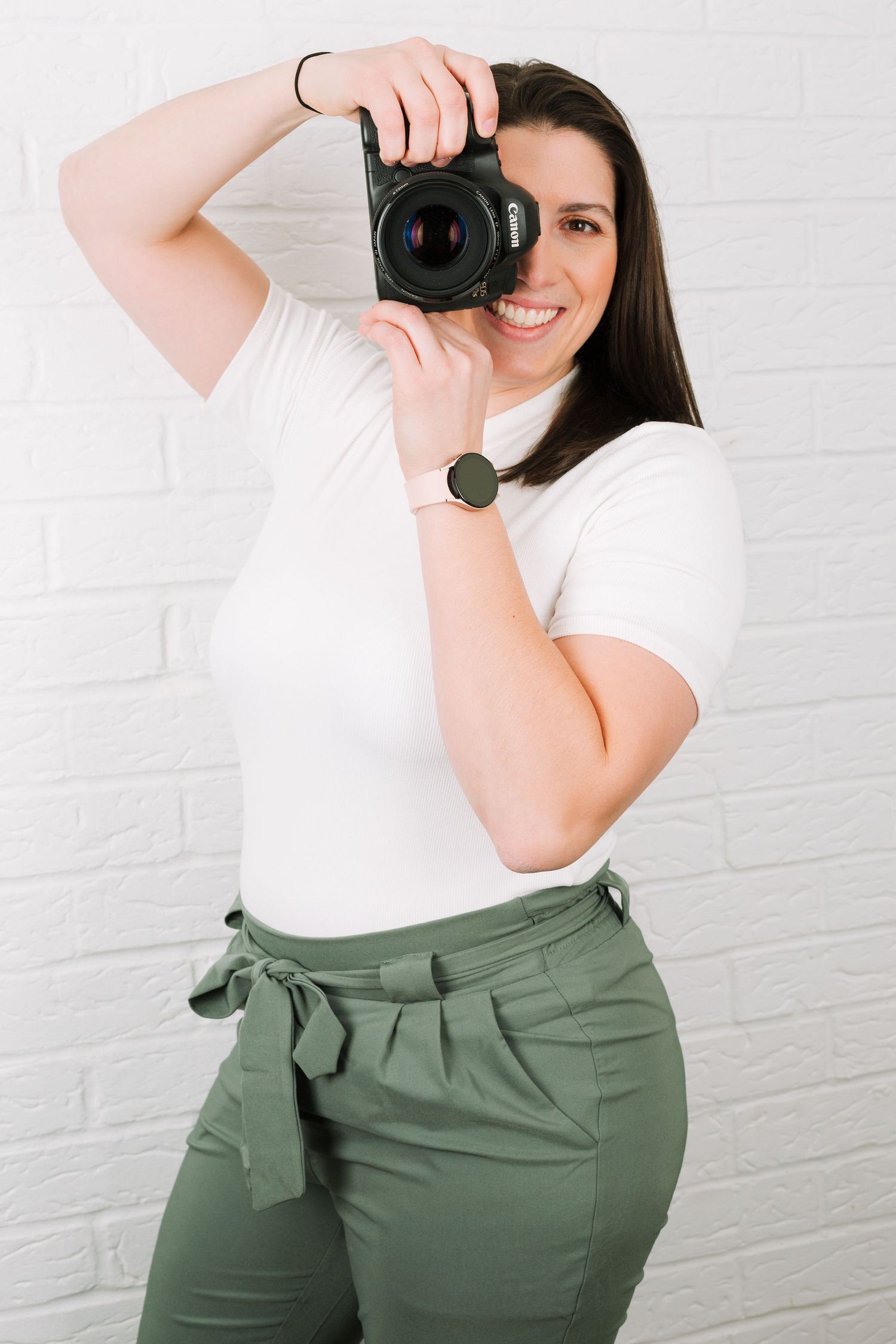 Photographer wearing white top and olive pants holds professional camera in front of white brick wall.