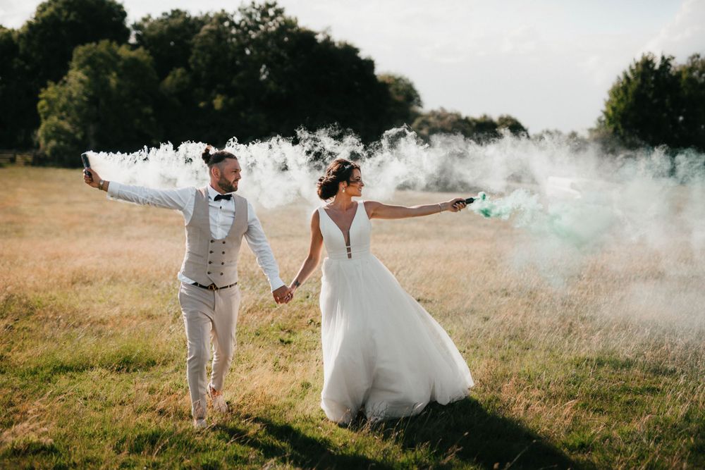 A couple celebrates with colorful smoke bombs in a sunny field during their wedding photoshoot.