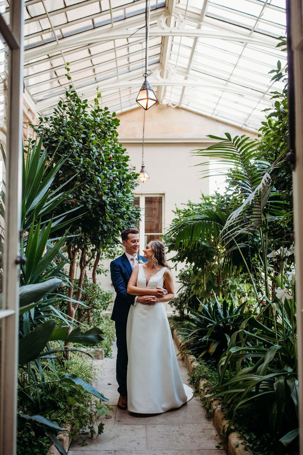 A wedding couple poses in a greenhouse filled with tropical plants and natural light.