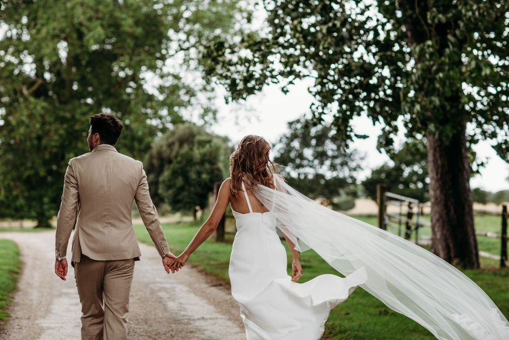 Couple walking down tree-lined path with flowing wedding veil.