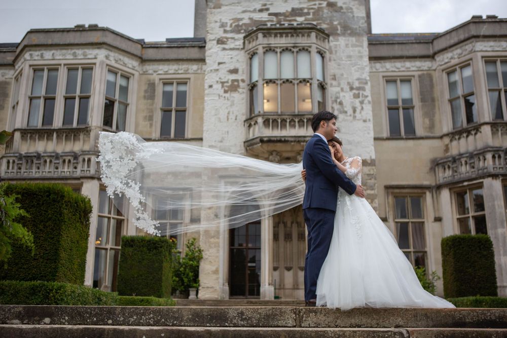 A wedding couple embraces as a veil flows in the wind outside a grand historic mansion with ornate architecture.