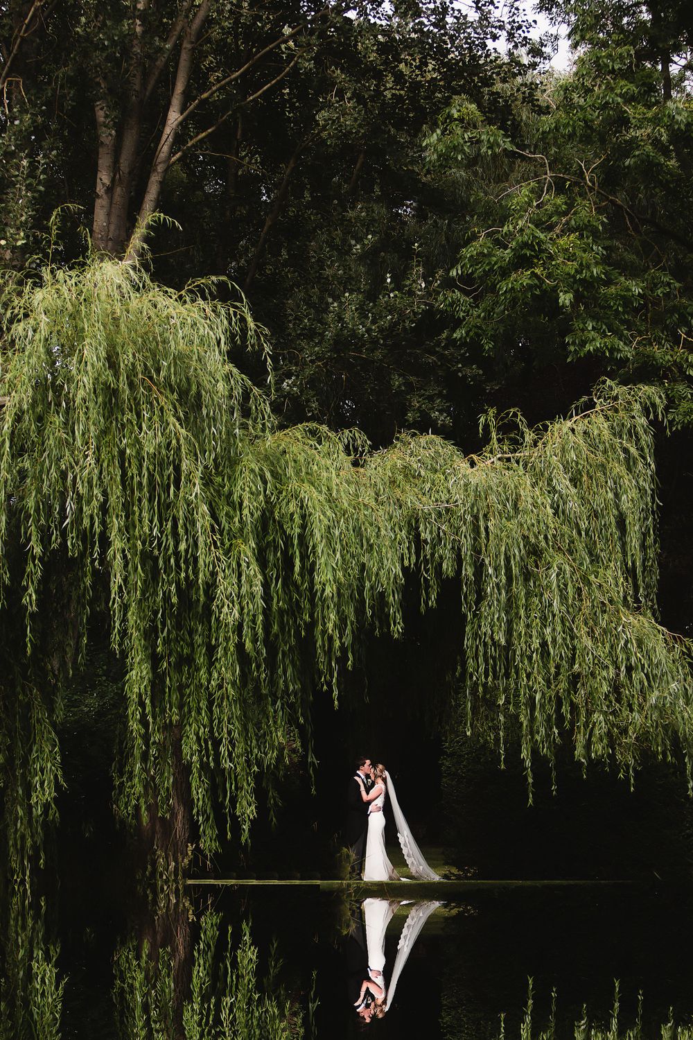 Dramatic wedding portrait under weeping willow trees reflecting in still water.