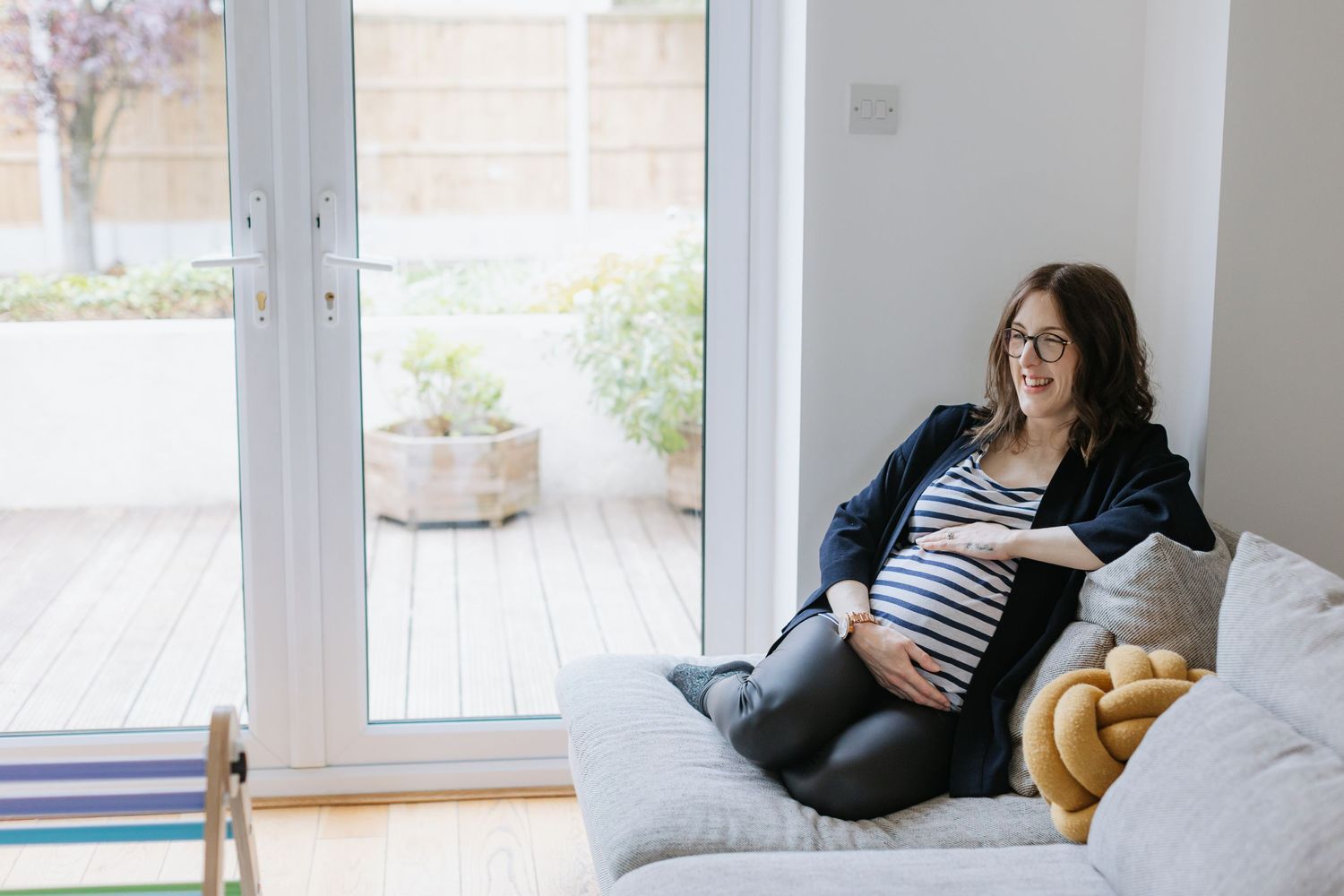 A pregnant woman relaxes on a cozy gray couch by large windows overlooking a patio.