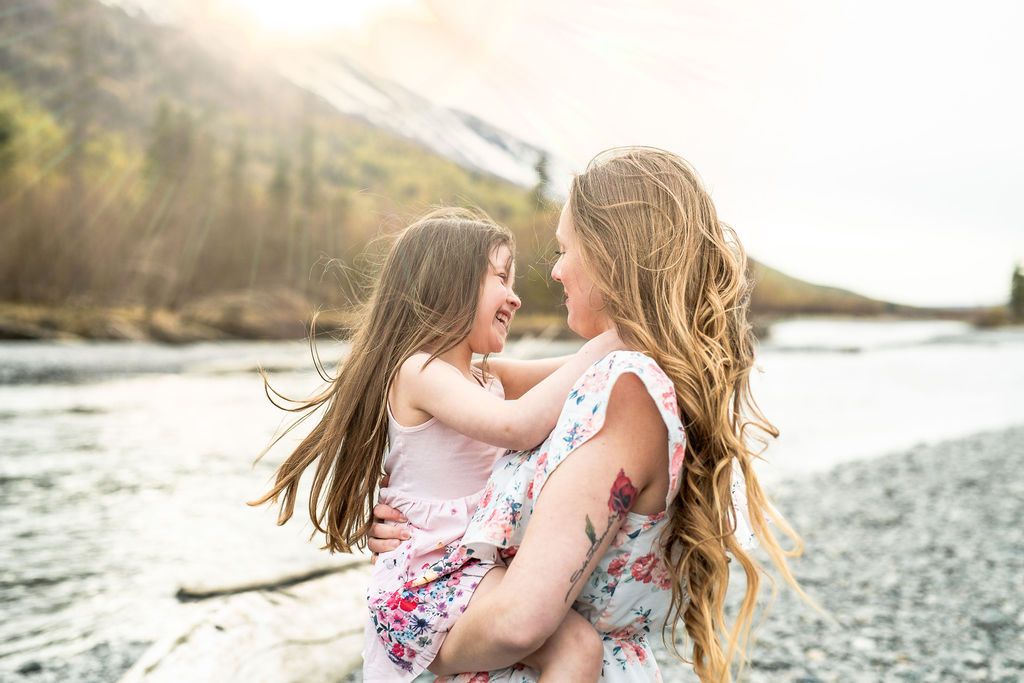 Mother and daughter sharing tender moment by mountain river in floral dresses.