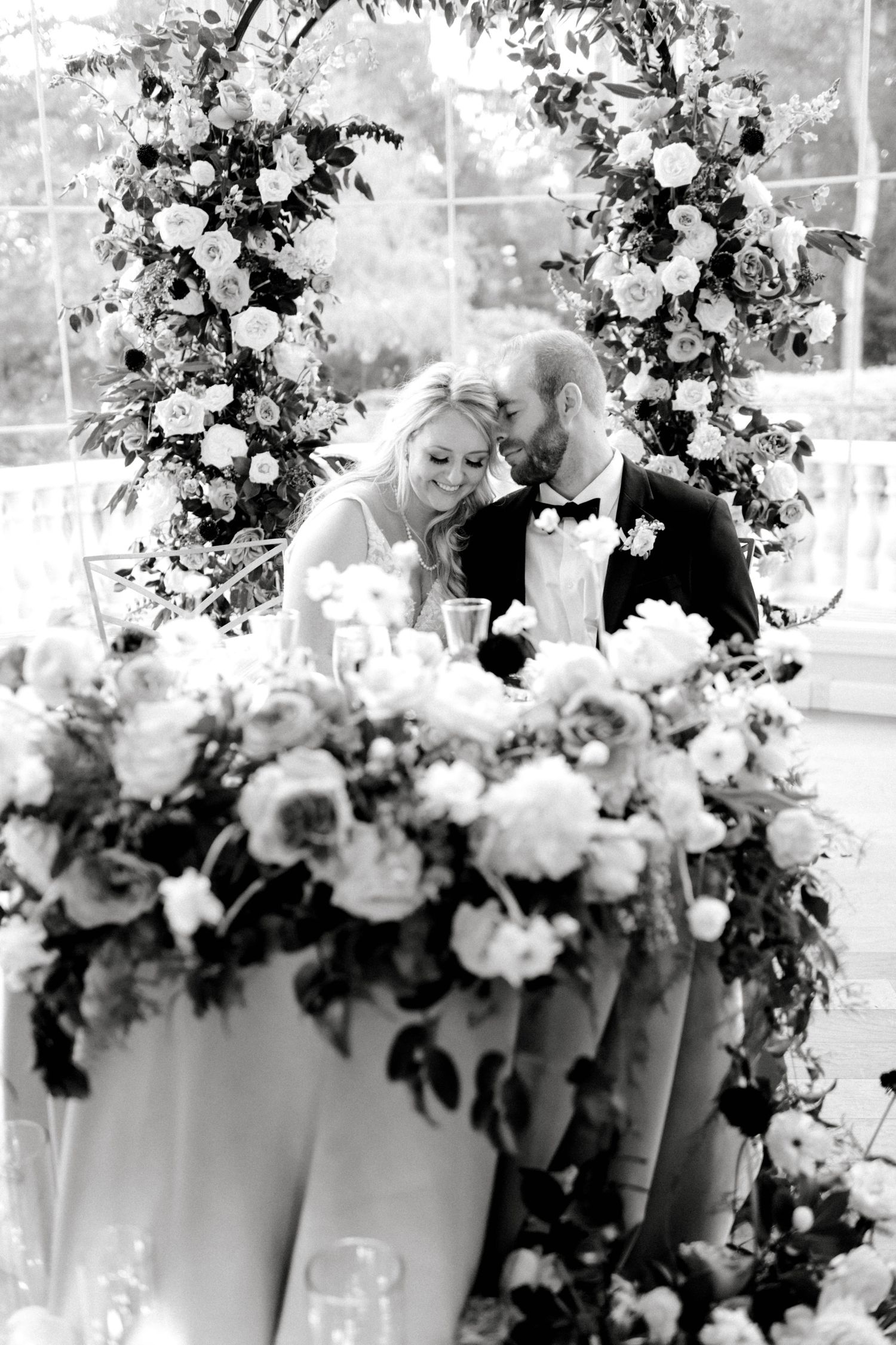 A romantic black and white photo of a couple at their wedding reception surrounded by lavish floral arrangements.